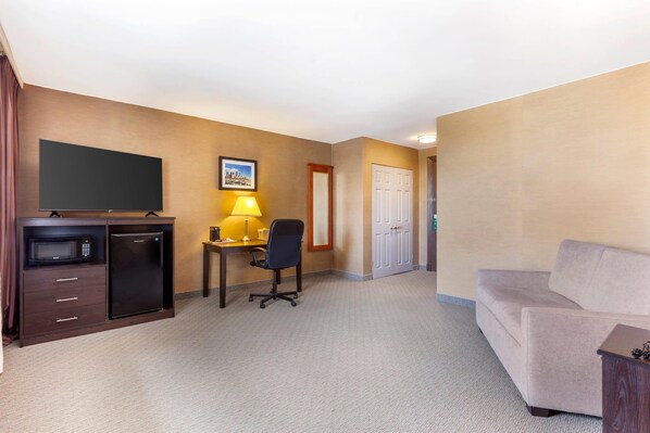 A hotel room with a flat-screen TV, a refrigerator, a desk with a lamp, a chair, and a sofa.