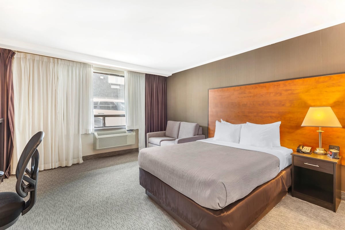 A hotel room with a large bed, a desk with a chair, a nightstand with a lamp, and a window with curtains.
