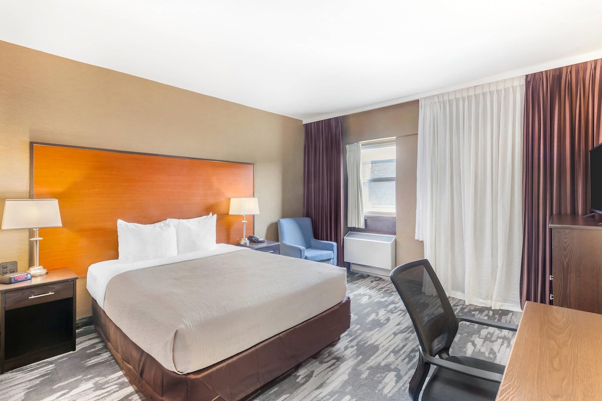 A hotel room with a large bed, a desk, a chair, a TV, and a window with curtains.
