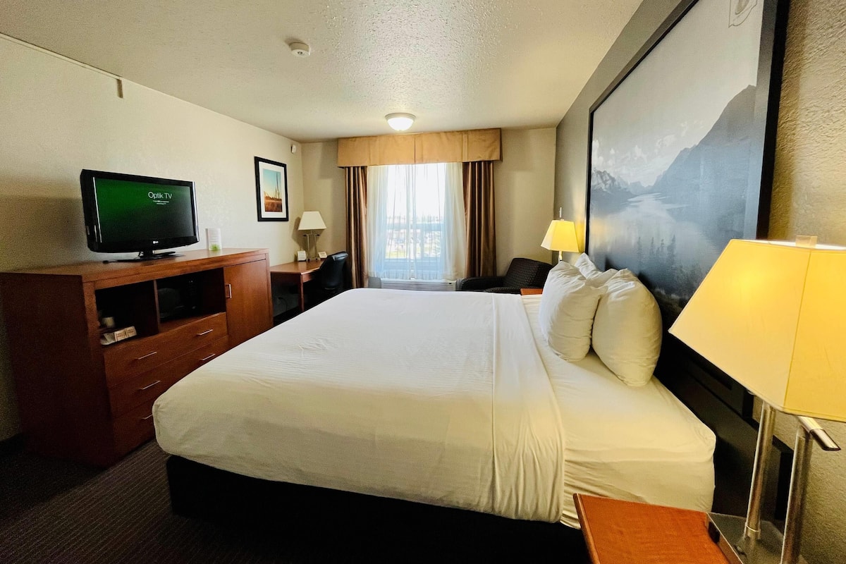 A hotel room with a large bed, a TV on a wooden cabinet, a desk with a chair, a lamp, and a painting on the wall.