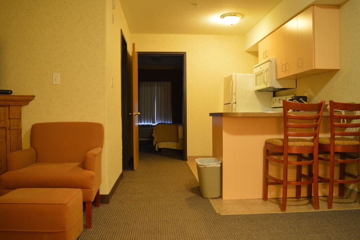 A compact hotel room with a kitchenette, a wooden chair, a small ottoman, and a trash bin.