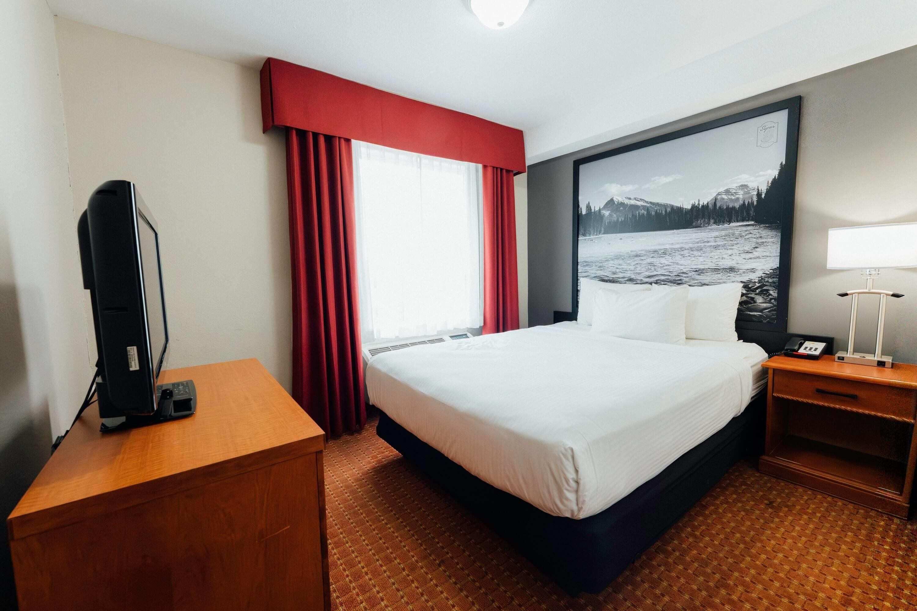 A hotel room with a bed, a desk, a TV, a lamp, and a large window with red curtains.