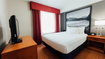 A hotel room with a bed, a desk, a TV, a lamp, and a large window with red curtains.