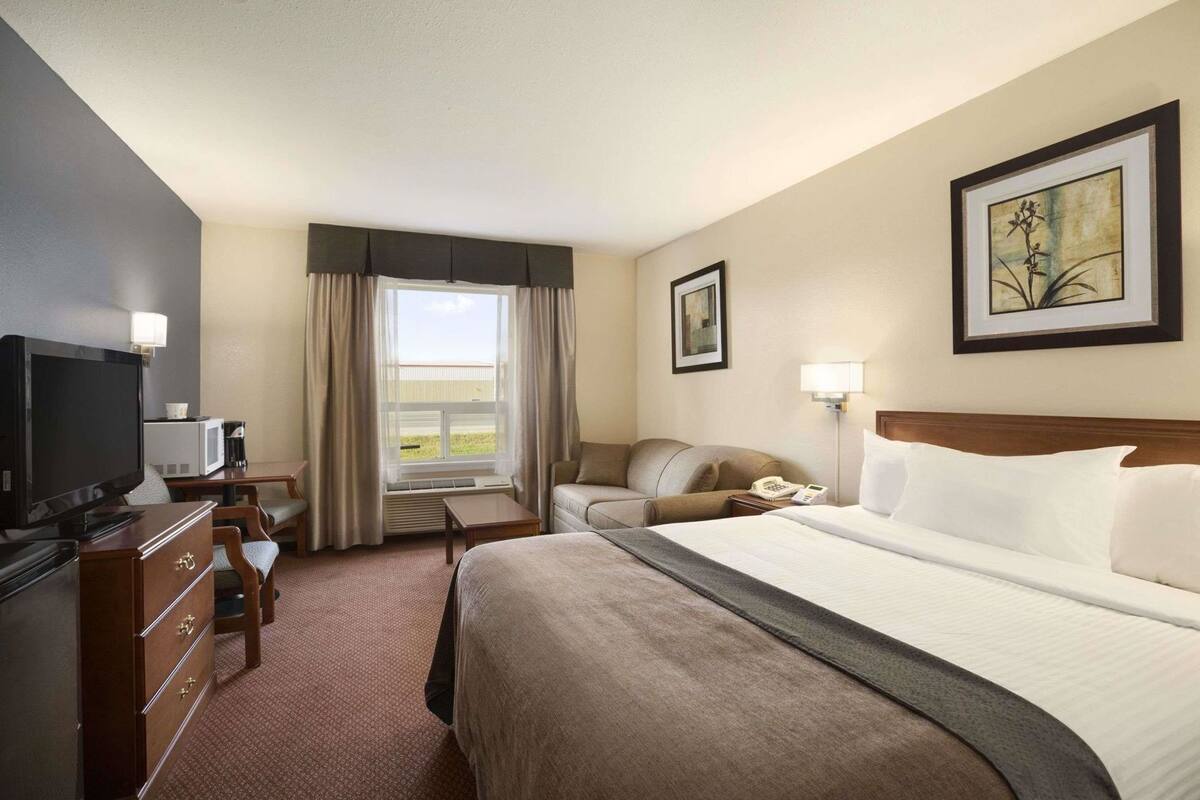 A hotel room with a bed, a television, a desk with a chair, a window with curtains, and a painting on the wall.