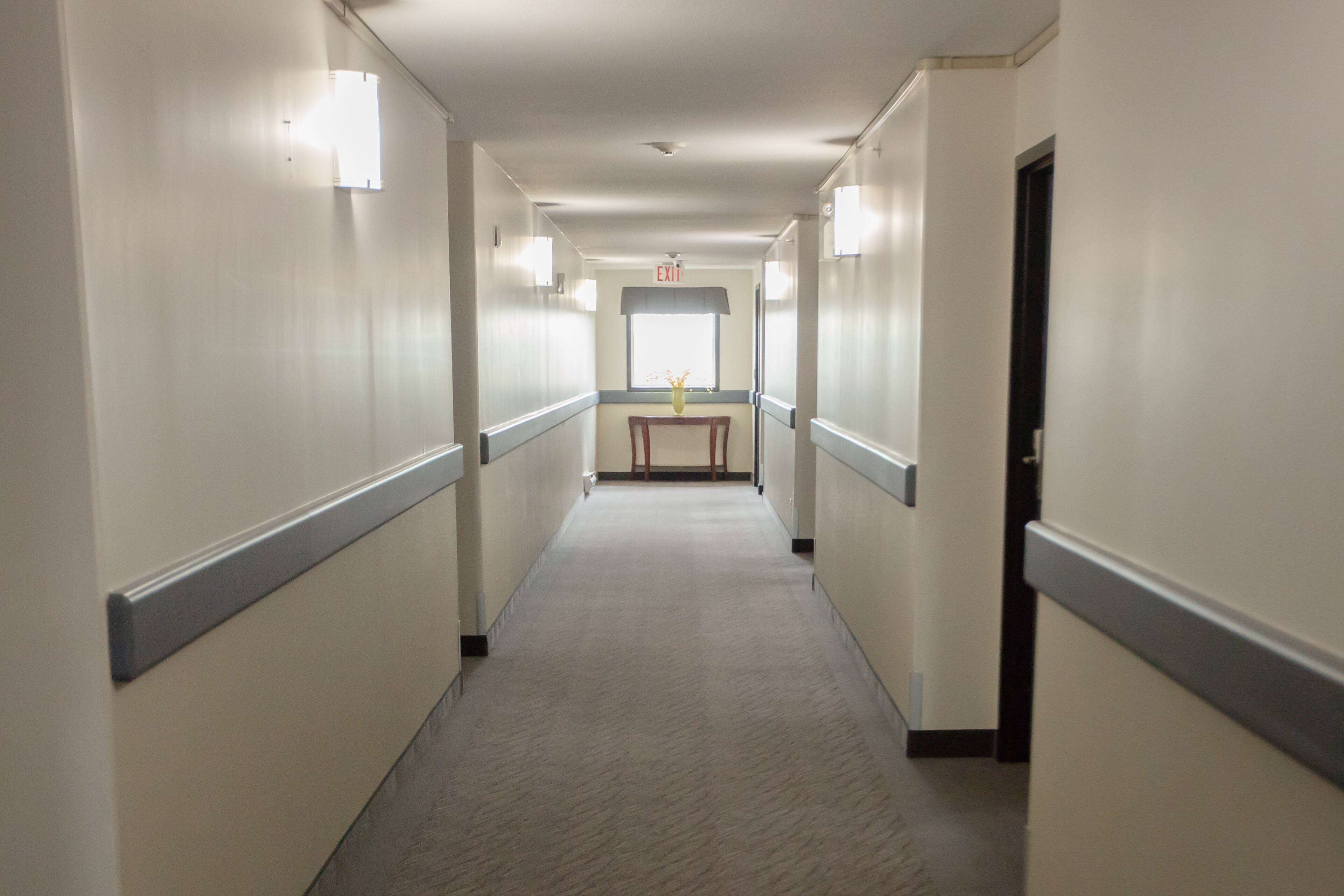 A long, narrow hotel corridor with a central table and chairs, light fixtures along the walls, and an exit sign above a window.