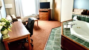 A hotel room with a wooden desk, a chair, a TV, a sofa, a large window with curtains, a bathroom with a bathtub, and a mirror. - Super 8 by Wyndham Whitecourt (Whitecourt)