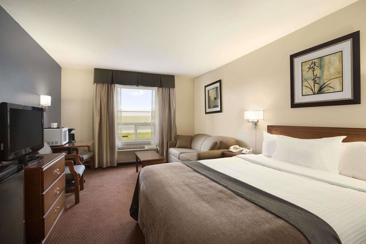 A hotel room with a bed, a television, a desk with a chair, a window with curtains, and a painting on the wall.