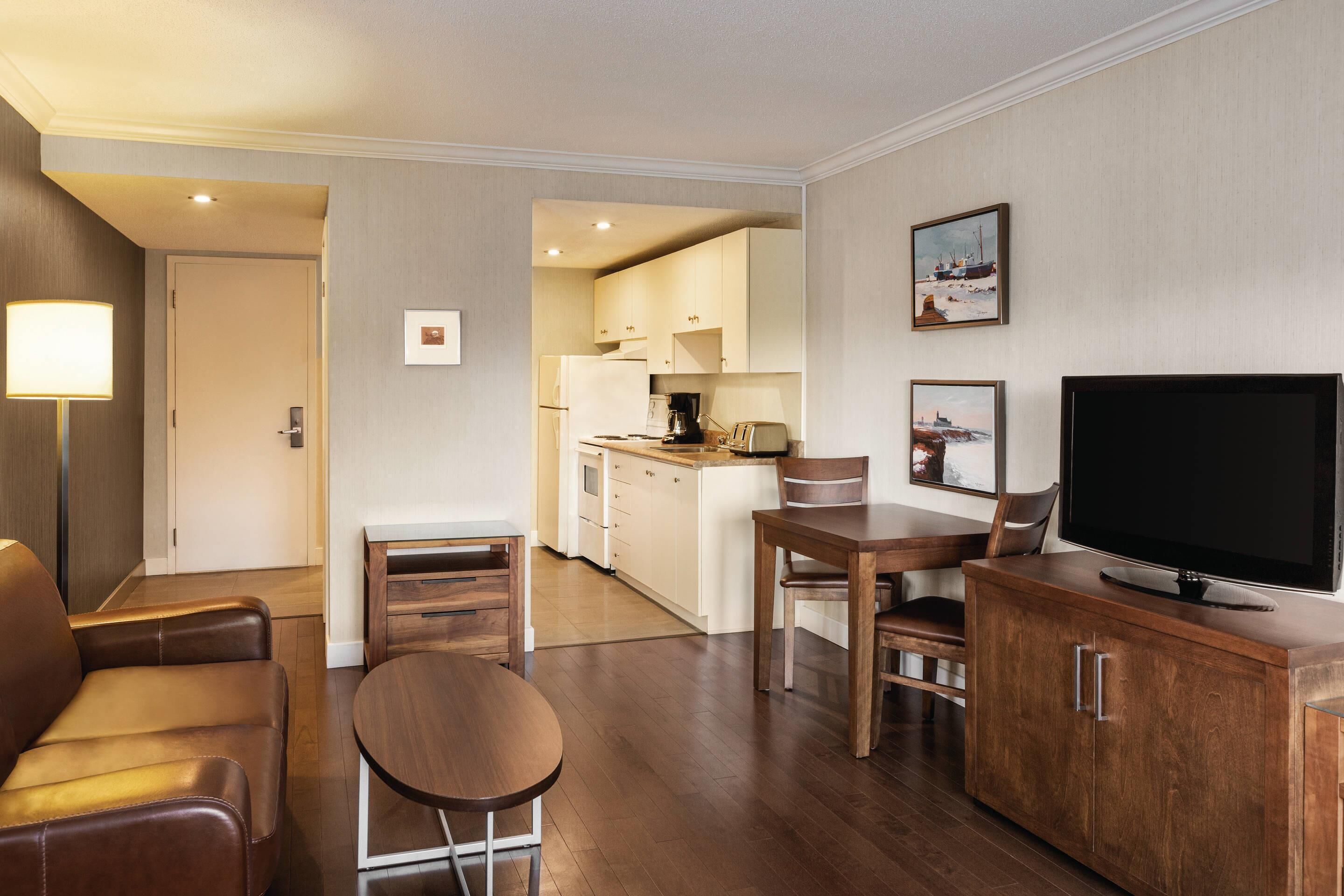 A modern hotel room with a living area, a dining table, a TV, and a kitchen in the background.