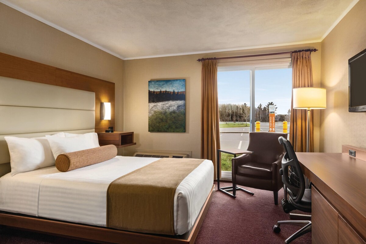 A hotel room with a bed, desk, chair, lamp, painting, and a view of the outside.