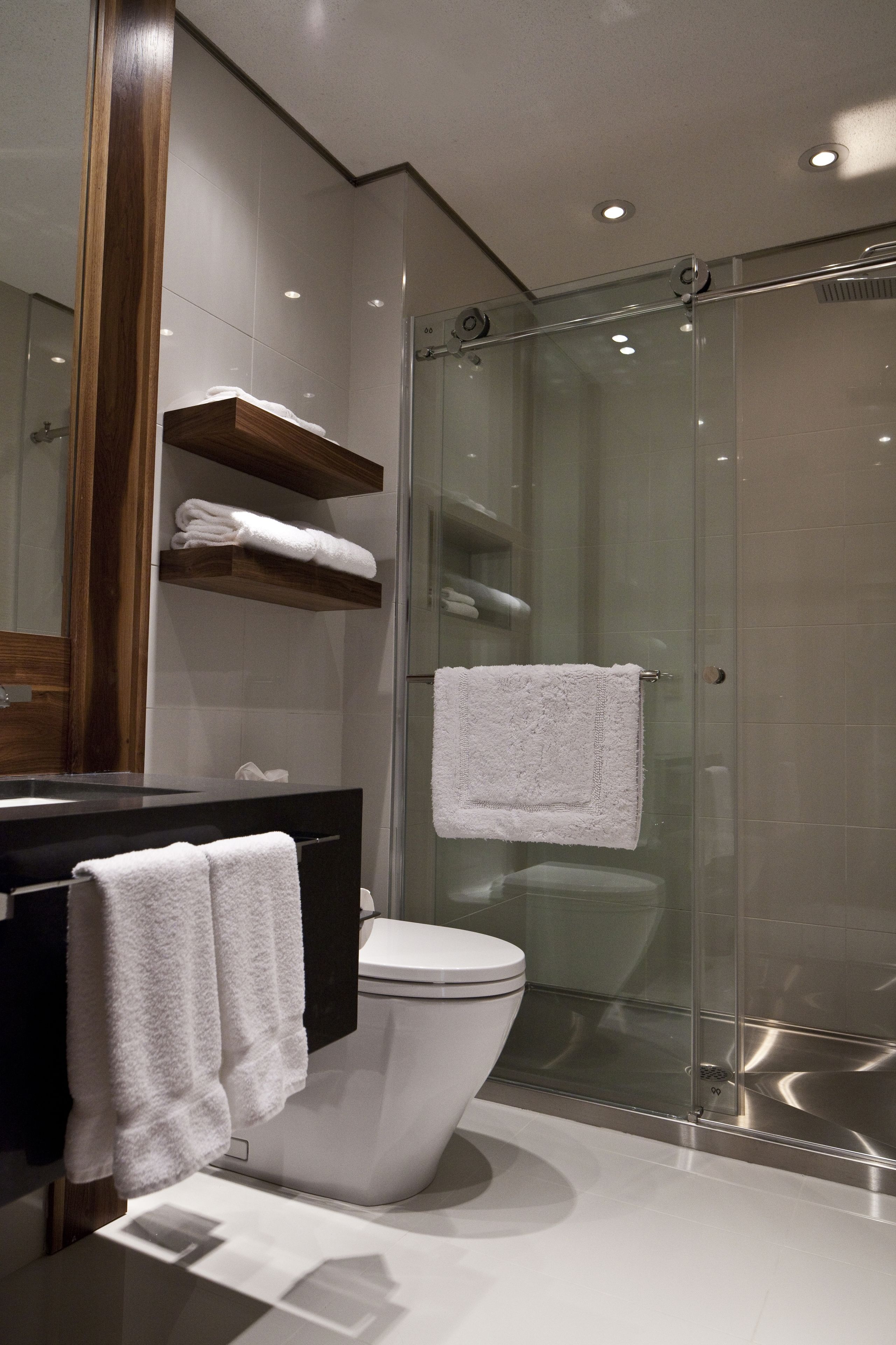 Un baño moderno con ducha envuelta en vidrio, inodoro blanco y tocador de madera con repisas para toallas.
