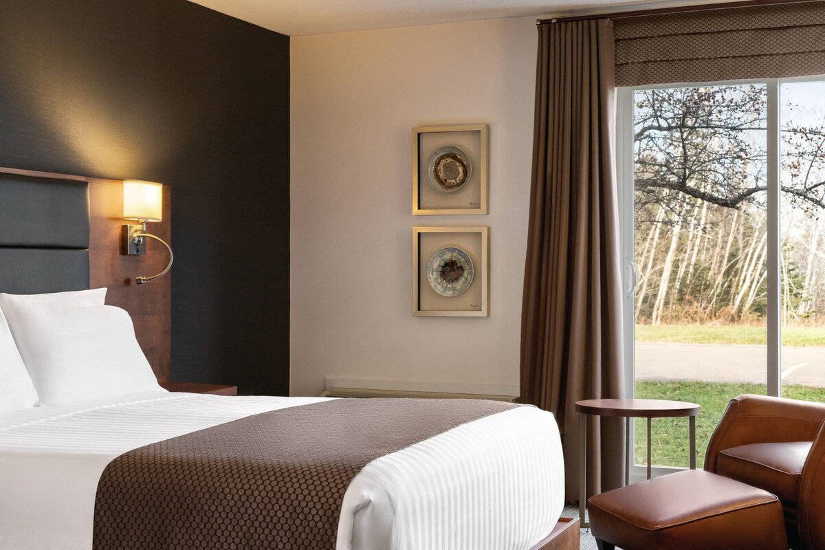 A hotel room with a bed, a small table, a chair, and two framed pictures on the wall.
