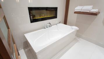 A modern bathroom with a large white bathtub, a mirror with a black frame, and a wooden shelf with folded towels.