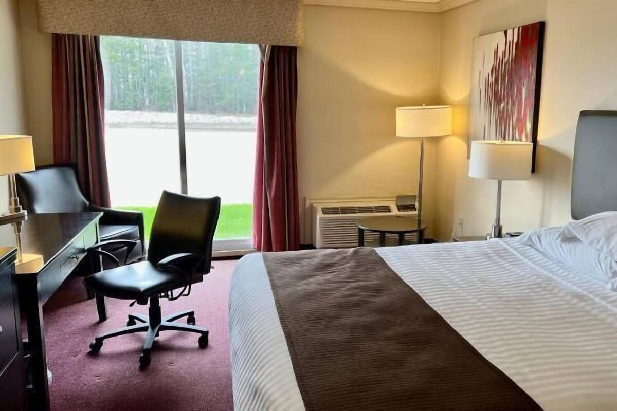 A hotel room with a bed, desk, chair, two lamps, and an air conditioning unit.