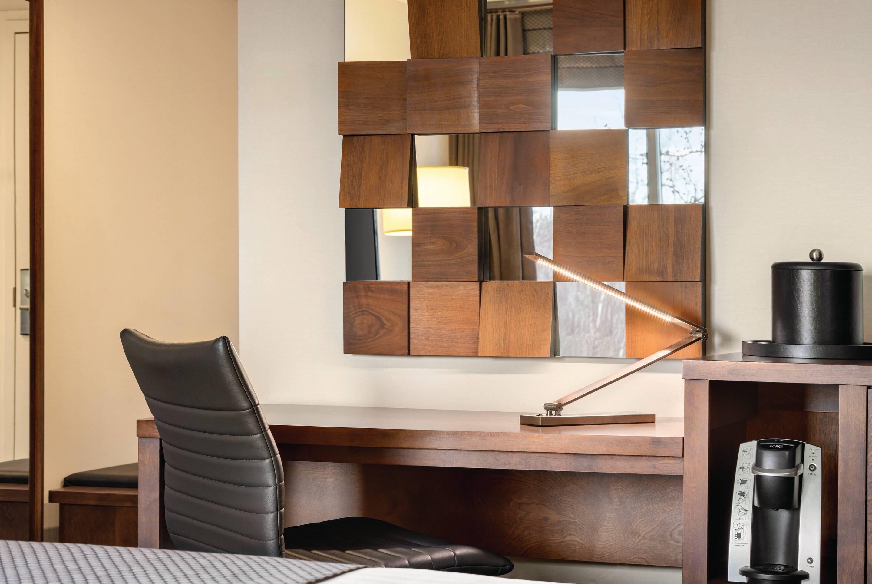 A hotel room with a wooden desk, a black leather chair, a modern lamp, and a coffee machine.