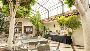 A spacious indoor dining area with glass tables and wicker chairs, surrounded by large trees and a high ceiling with glass panels. - Clocktower Inn Ventura (Ventura)