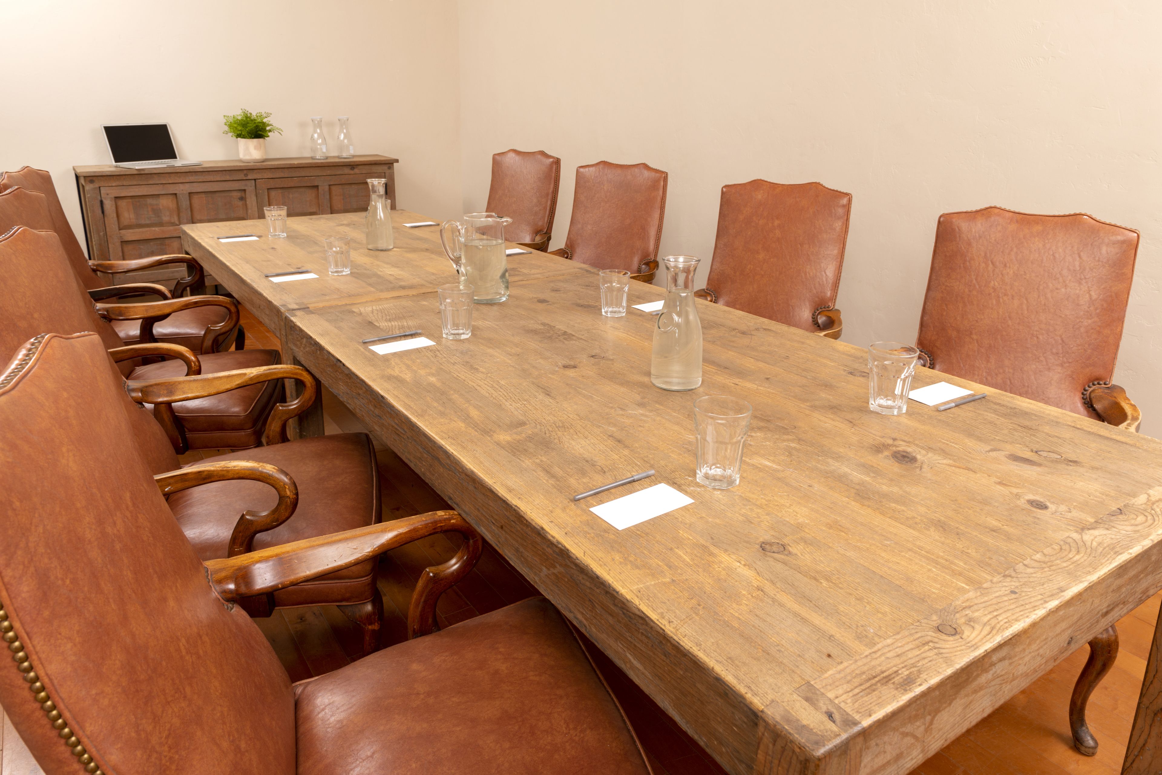 A conference room with a long wooden table, leather chairs, and a laptop on a cabinet.