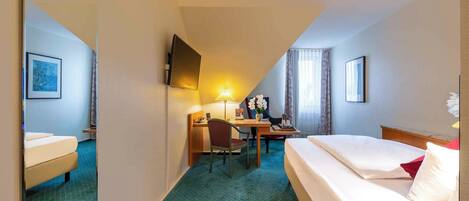 Habitación de hotel con cama, escritorio con silla, televisor montado en la pared y ventana con cortinas.