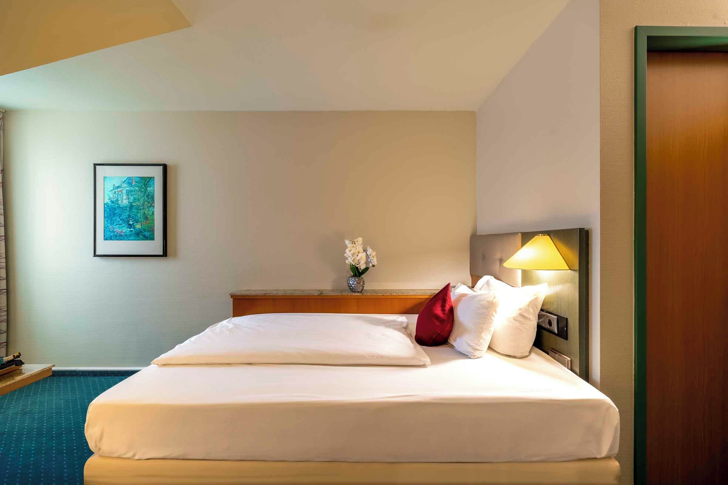 A hotel room with a bed, a nightstand, a lamp, a framed picture on the wall, and a vase with flowers.
