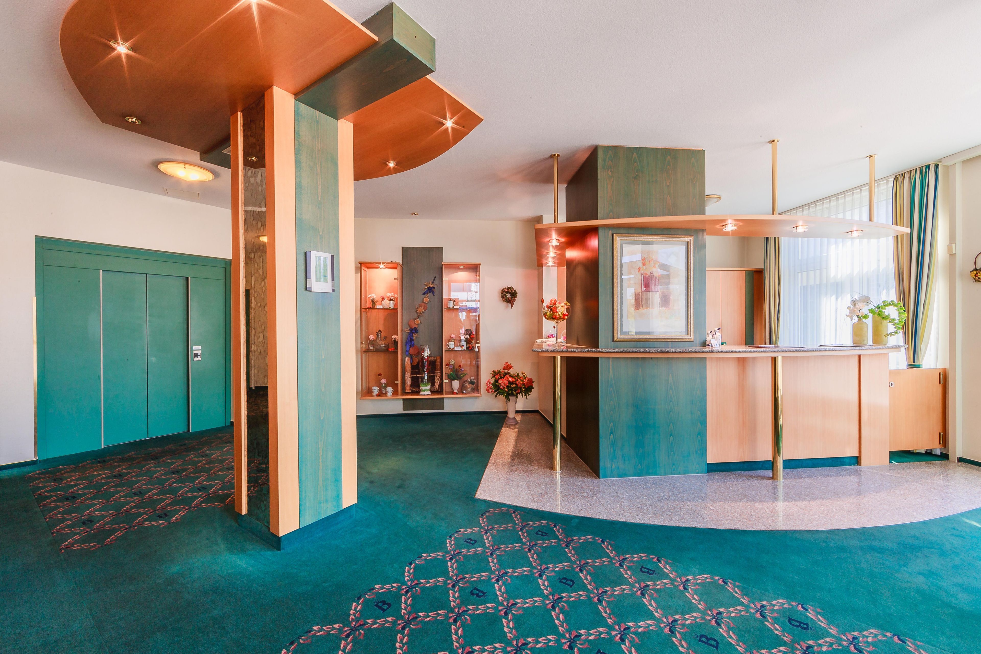 A hotel lobby with a teal carpet, a reception desk, and a wall-mounted TV.