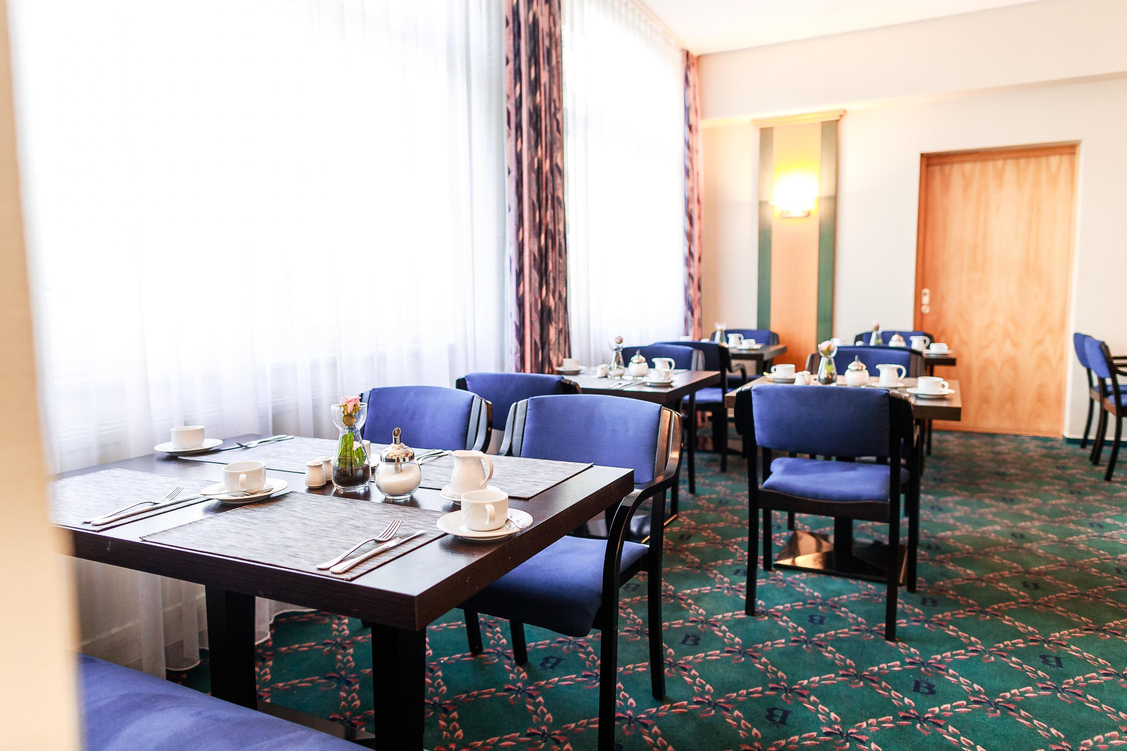 Comedor de hotel con mesas dispuestas para cuatro, sillas azules y alfombra estampada.