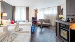 A hotel room with two beds, a sofa, a dining table, a TV, and a minibar.