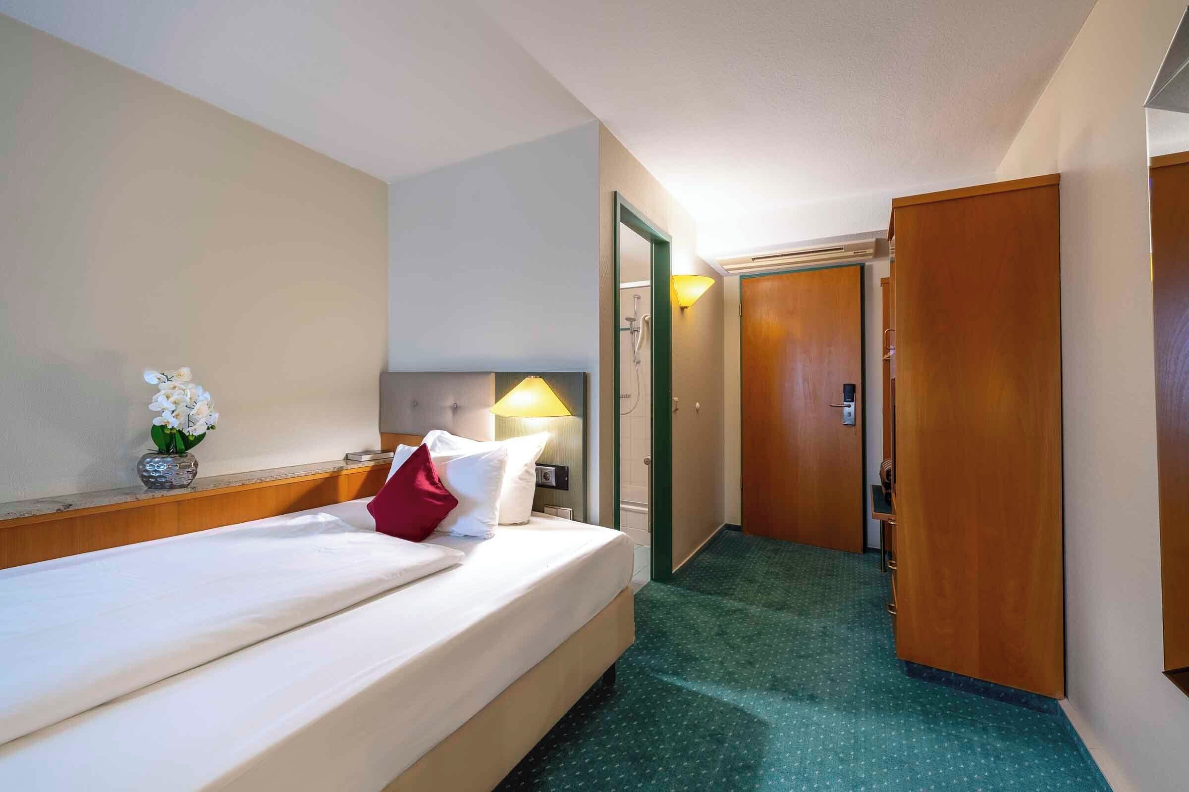 A hotel room with a bed, a nightstand, a lamp, a vase with flowers, and a door leading to another room.