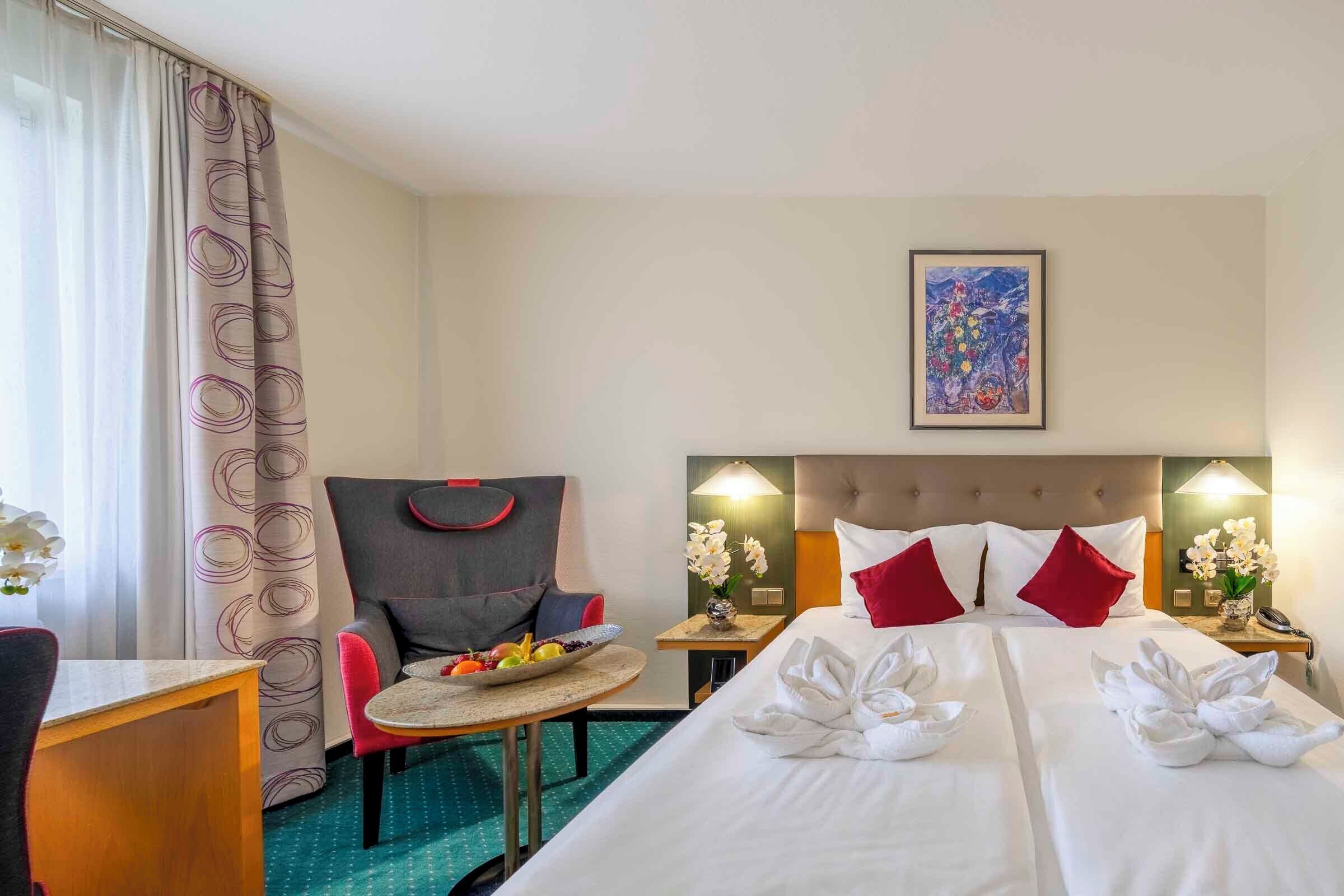 Habitació d'hotel amb dos llits, una taula de menjador amb fruita, una cadira i un capçal amb una làmpada.