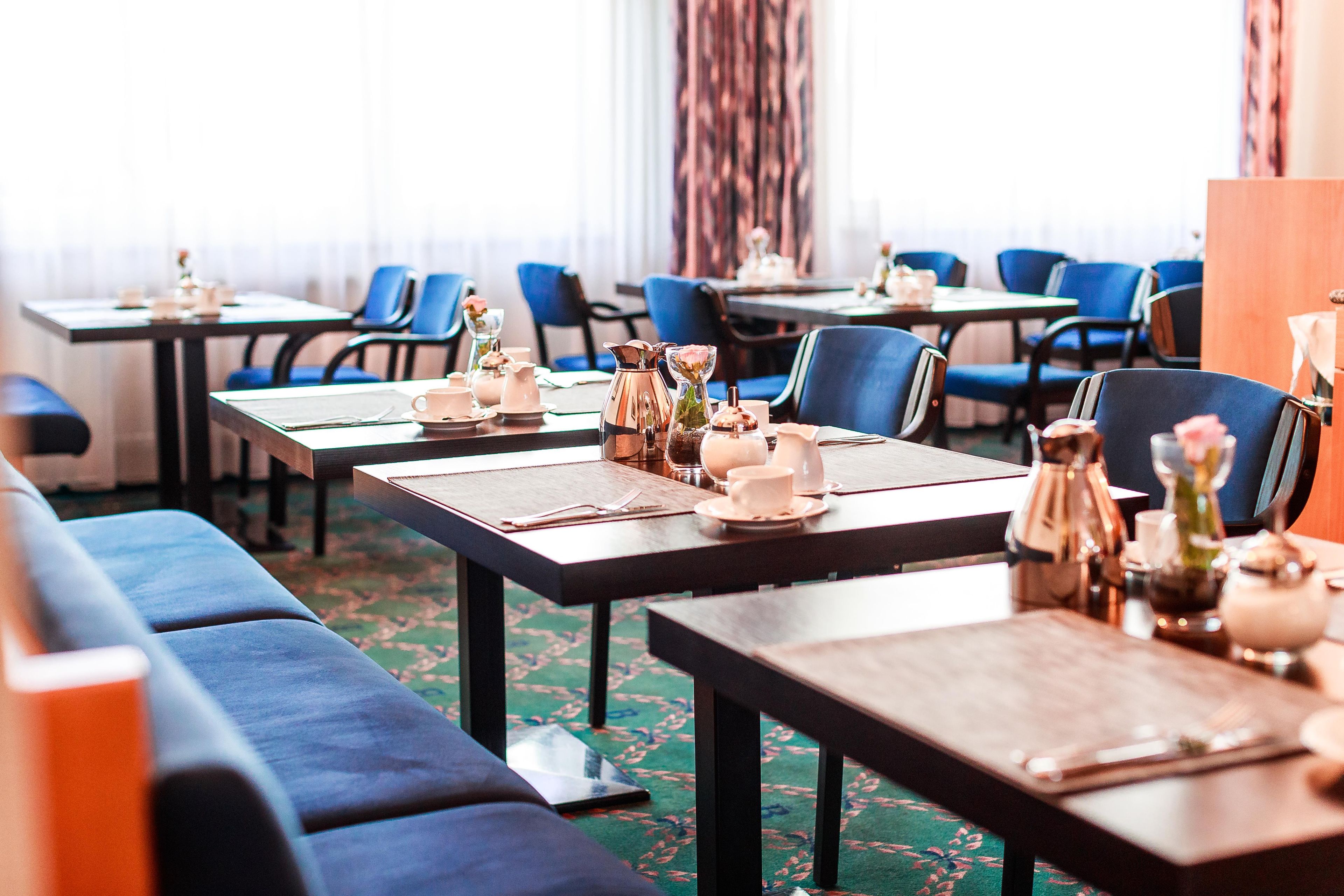 A hotel dining area with tables set for breakfast, blue upholstered chairs, and a carpeted floor.