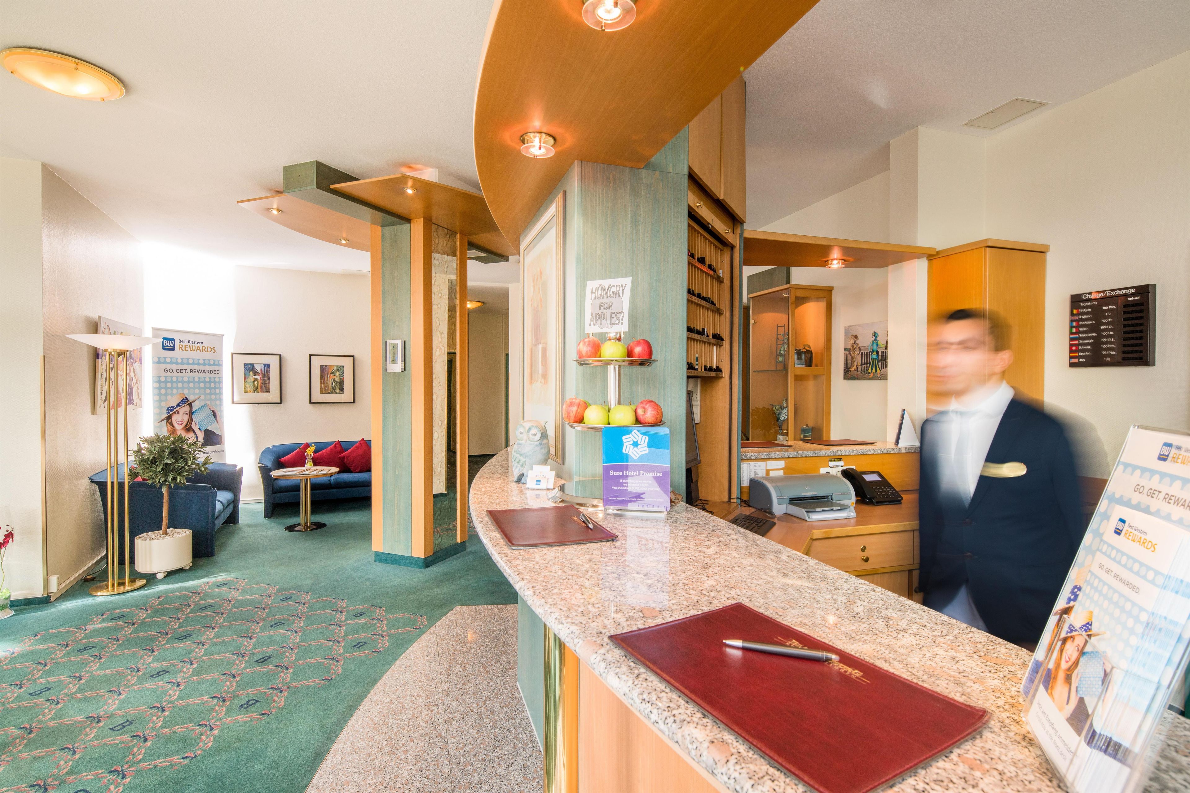 Un vestíbulo de hotel con una recepción, un sofá azul, obras de arte enmarcadas y un cartel que ofrece recompensas del hotel.