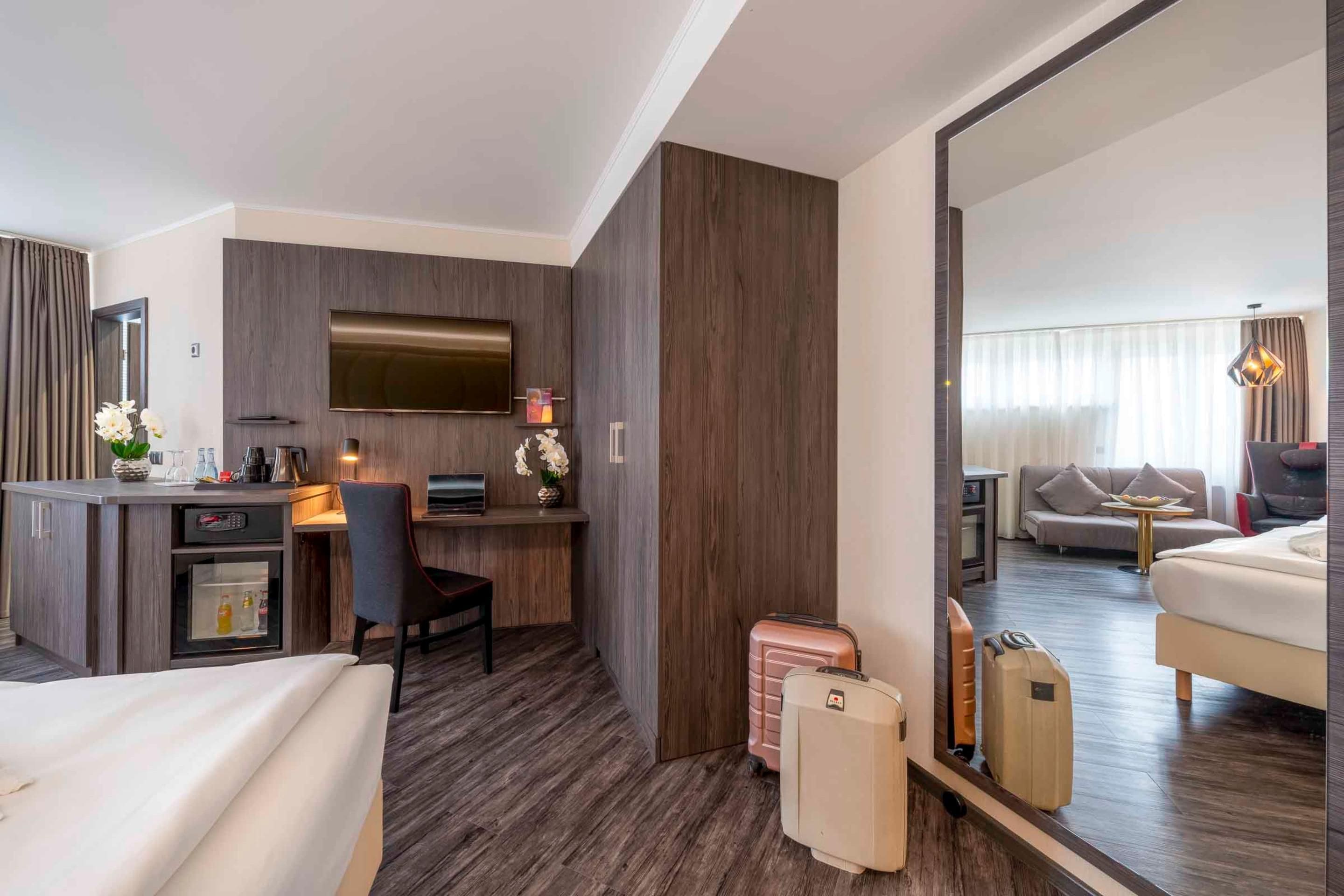 Habitación de hotel con una cama, un escritorio con computadora, un televisor y un espejo grande que refleja la habitación.