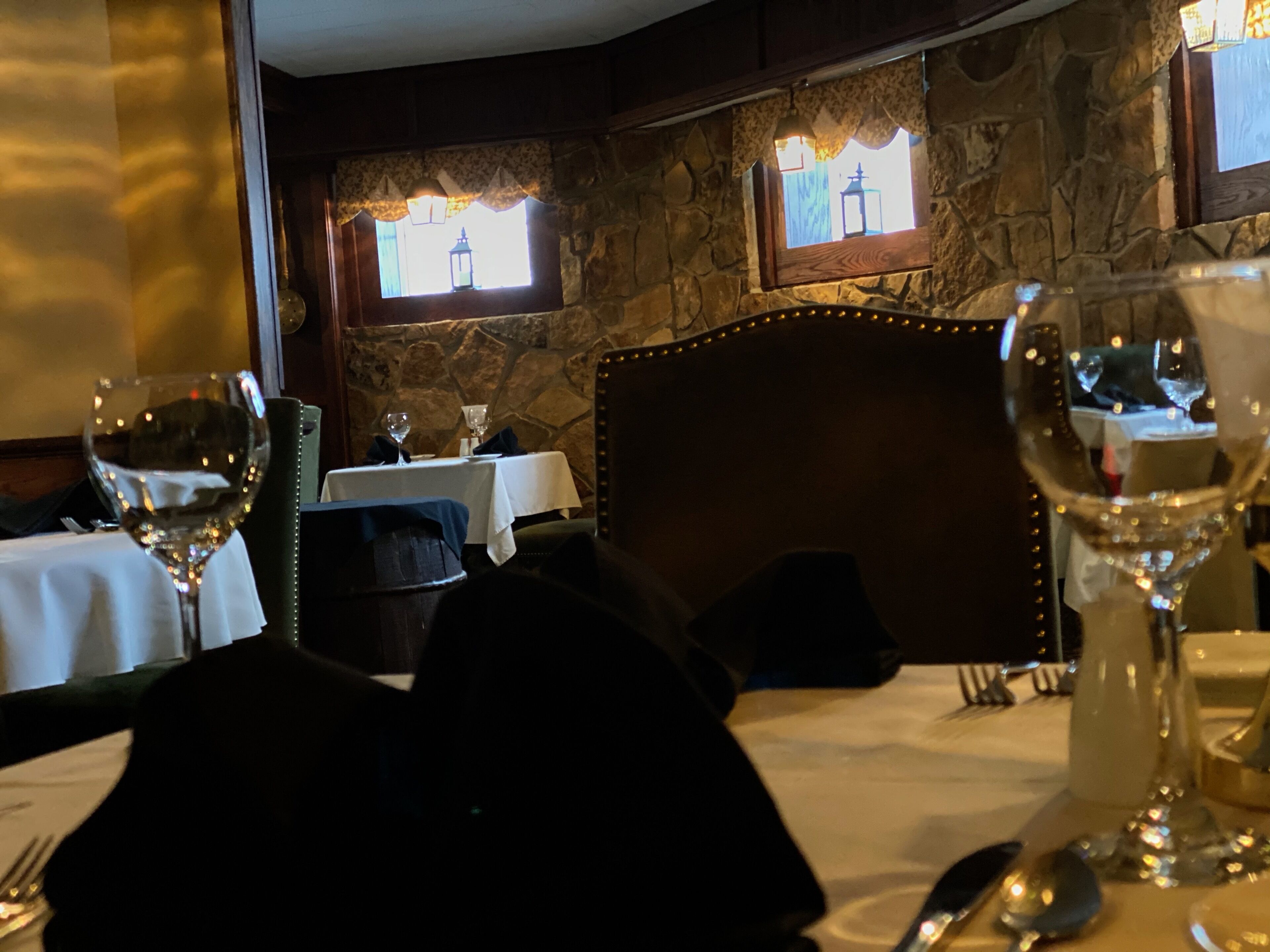Un comedor tenuemente iluminado con paredes de piedra, un cabecero de madera y una mesa para dos, con copas de vino y cubiertos.