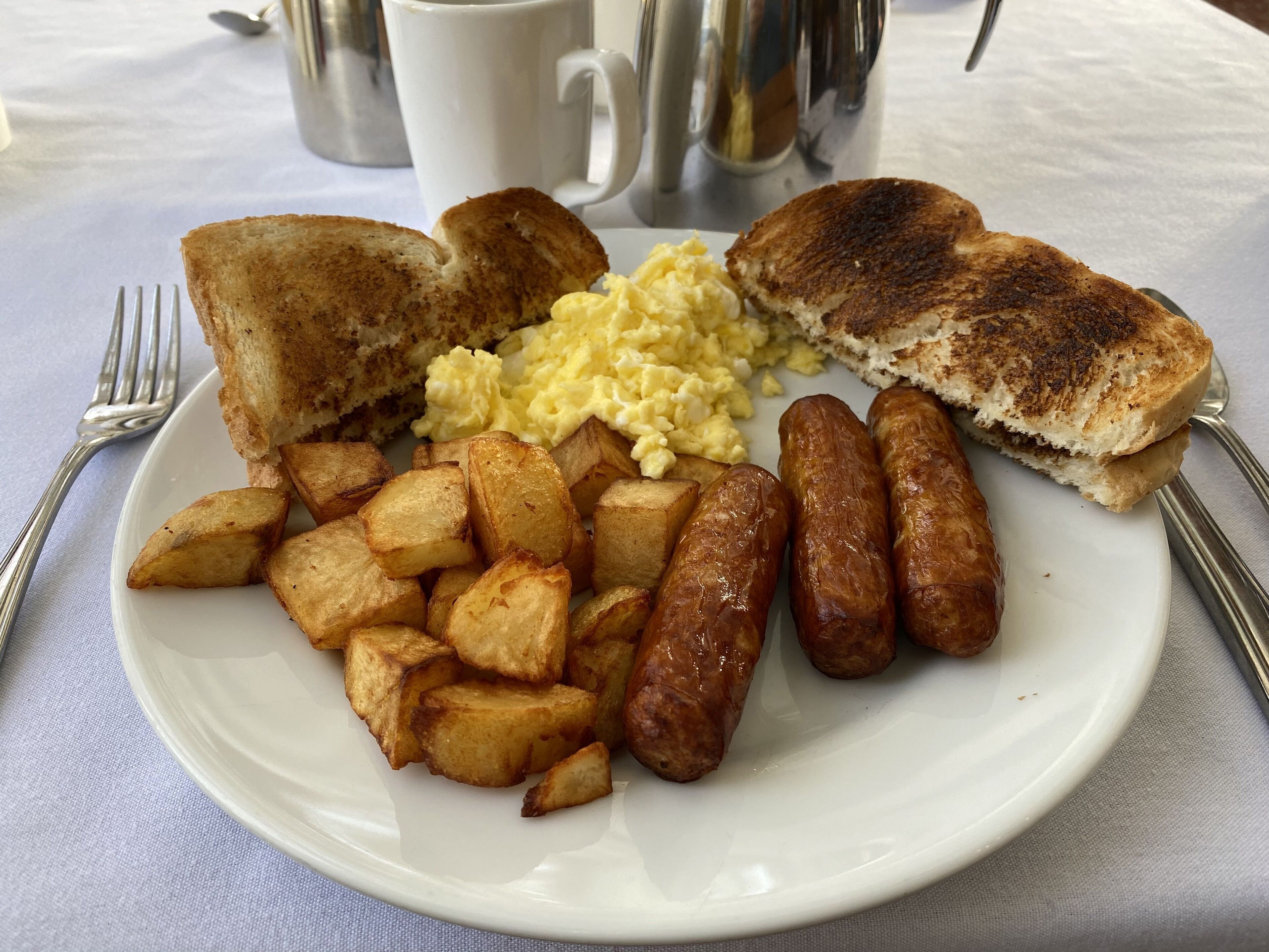 Un plato de comida para el desayuno con tostadas, huevos revueltos, salchichas y patatas asadas.