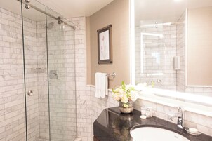 Un baño moderno con ducha acristalada, pared de azulejos de mármol, espejo grande y encimera negra con un florero.