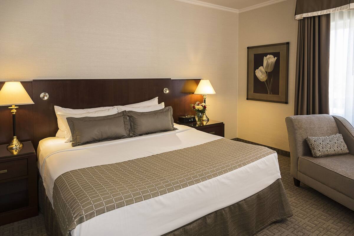 Hotellihuone, jossa on suuri sänky, kaksi yöpöytää valaisimineen, yöpöydän päällä oleva lamppu, sohva ja seinällä oleva kehystetty kuva.