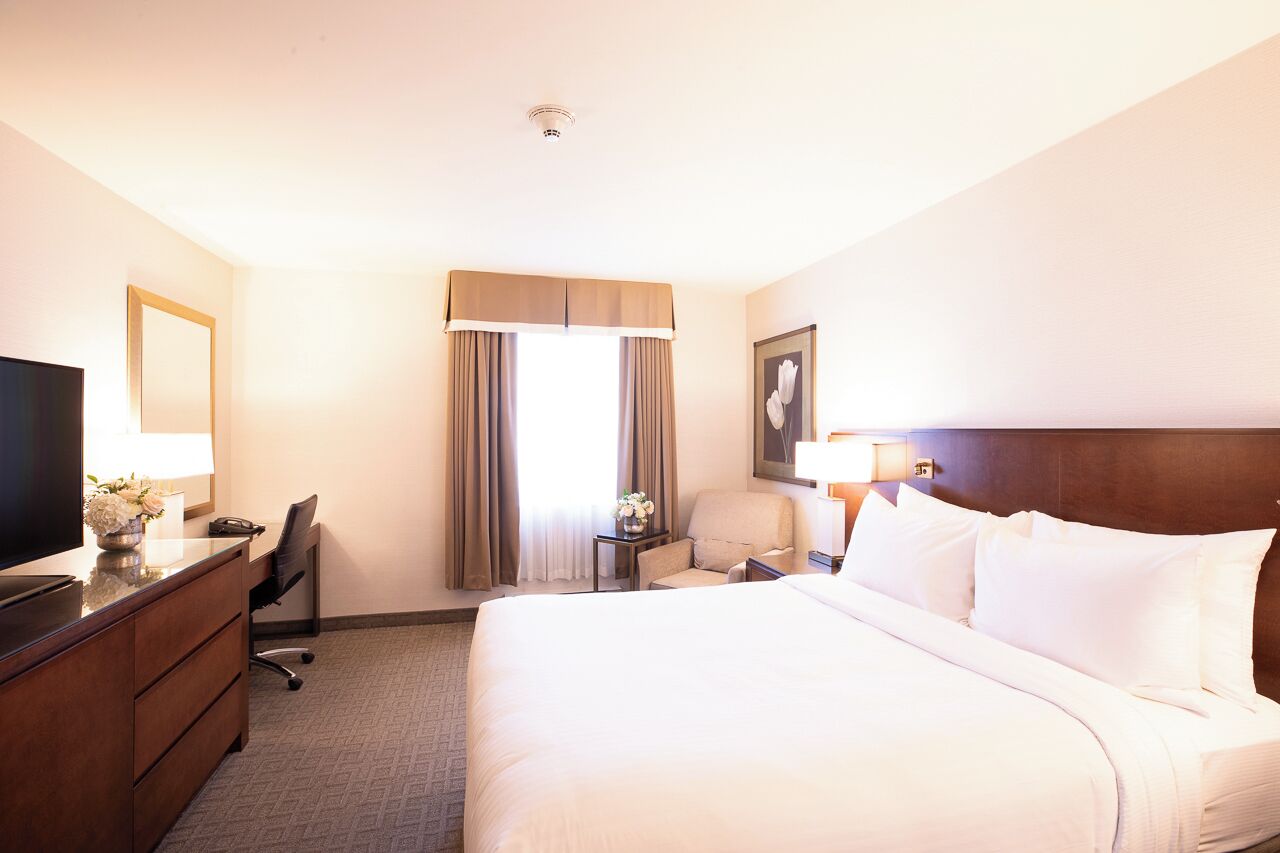Habitación de hotel con una cama grande, un escritorio con computadora, una silla, un televisor y una ventana con cortinas.
