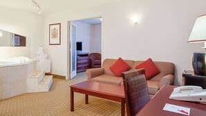 A hotel room with a bathroom, a sofa, a table, and a TV. - Ramada by Wyndham Red Deer Hotel and Suites (Red Deer)