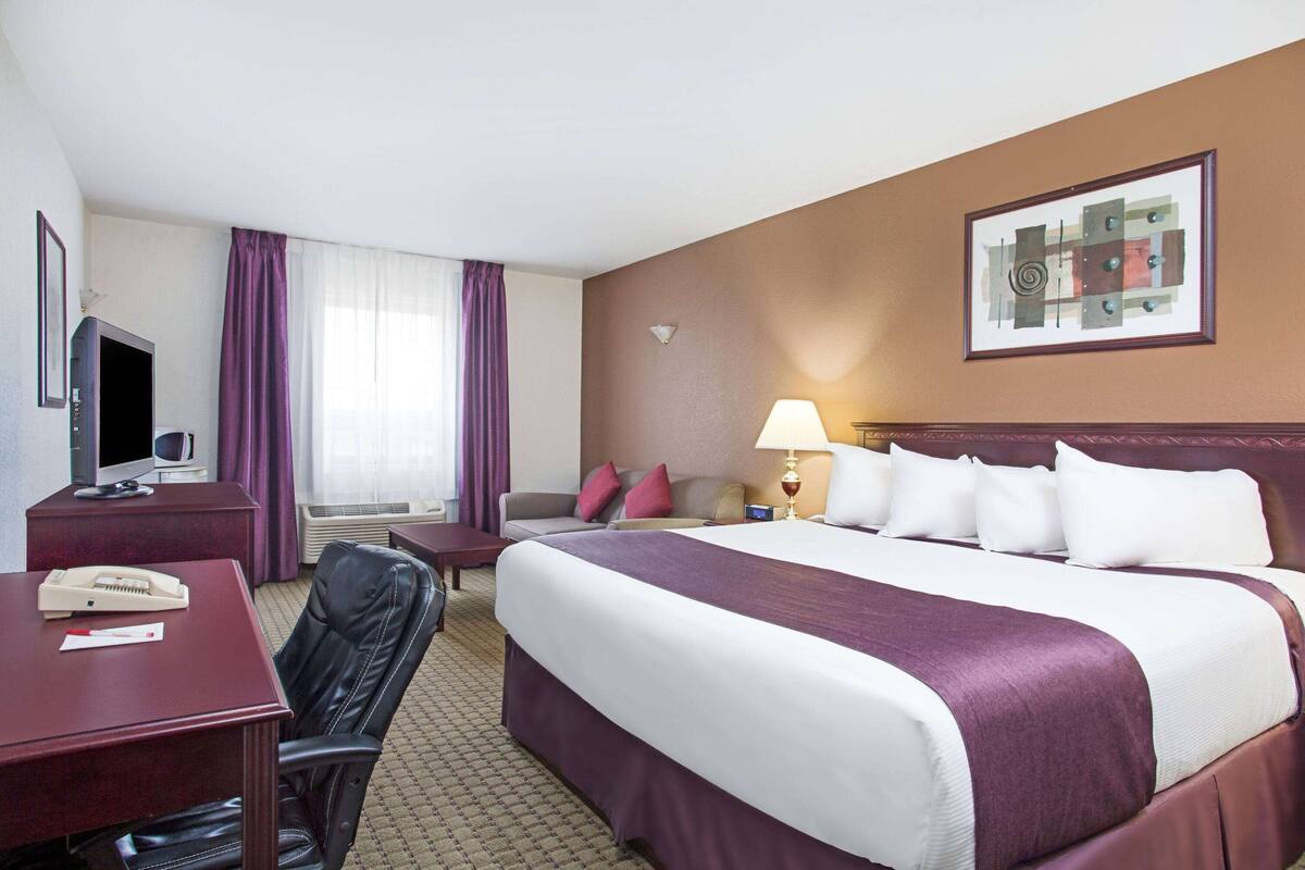 Une chambre d’hôtel avec un grand lit, un bureau avec un téléphone, une chaise, un téléviseur et une fenêtre avec des rideaux violets.