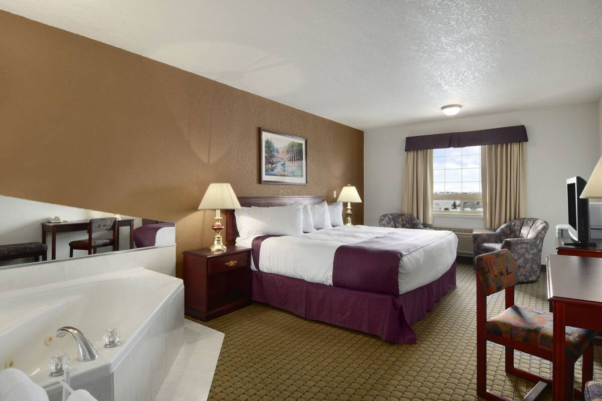 Une chambre d’hôtel avec un grand lit, un coin salon avec un fauteuil, un bureau, une télévision, une fenêtre avec des rideaux et une salle de bain avec une baignoire et un lavabo.