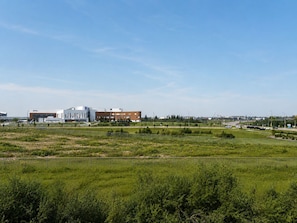 A large open field with a fence in the foreground, a road with vehicles in the background, and a building with multiple floors and a flat roof.