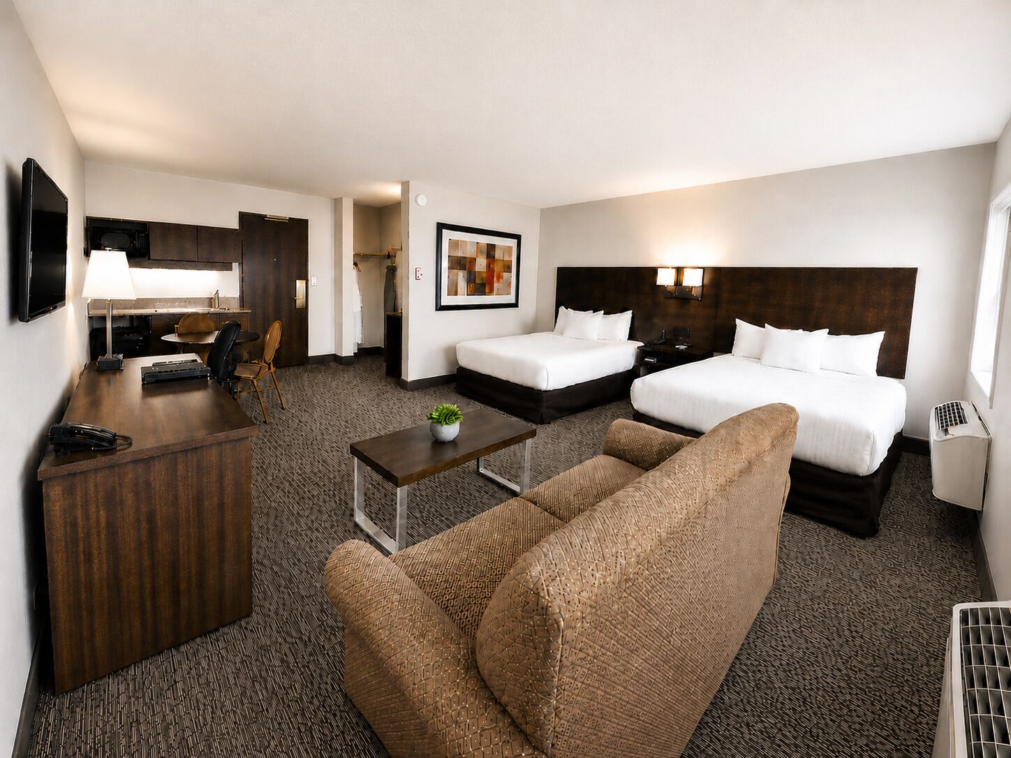 A hotel room with two beds, a desk, a chair, a sofa, a small table with a plant, and a picture on the wall.