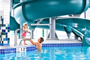 Uma piscina com escorrega aquático, uma criança e um adulto, e uma parede revestida com padrão azul e branco.