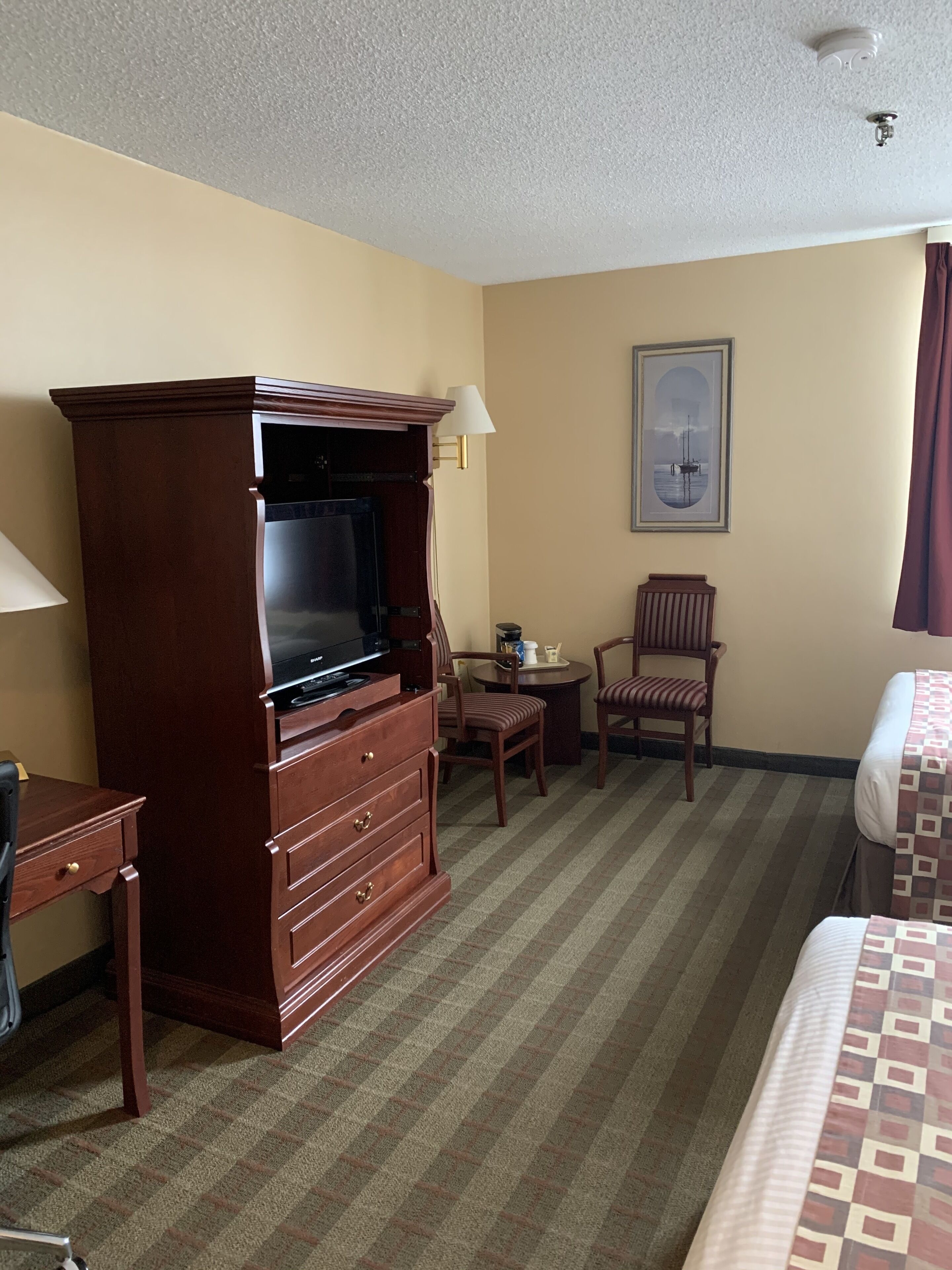Una habitación de hotel con un gran mueble de televisión de madera, una cama, un escritorio, una silla y una mesita con una lámpara.
