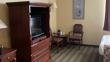 Una habitación de hotel con un gran mueble de televisión de madera, una cama, un escritorio, una silla y una mesita con una lámpara.