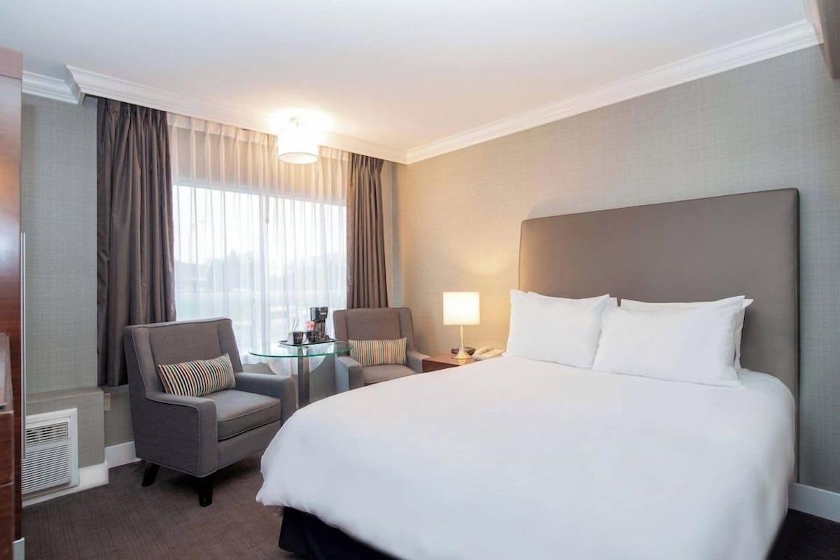A hotel room with a large bed, two armchairs, a small table with a lamp, and a window with curtains.