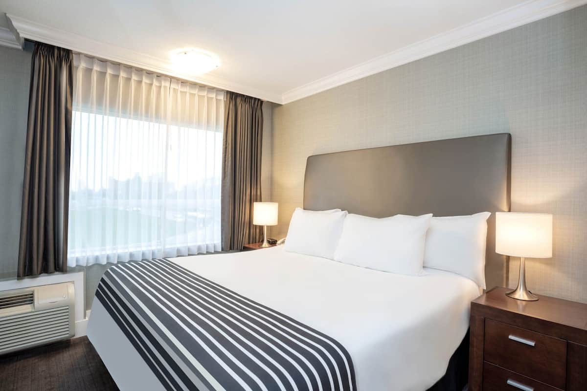 A hotel room with a large bed, two bedside lamps, a heater, and a view of the city.