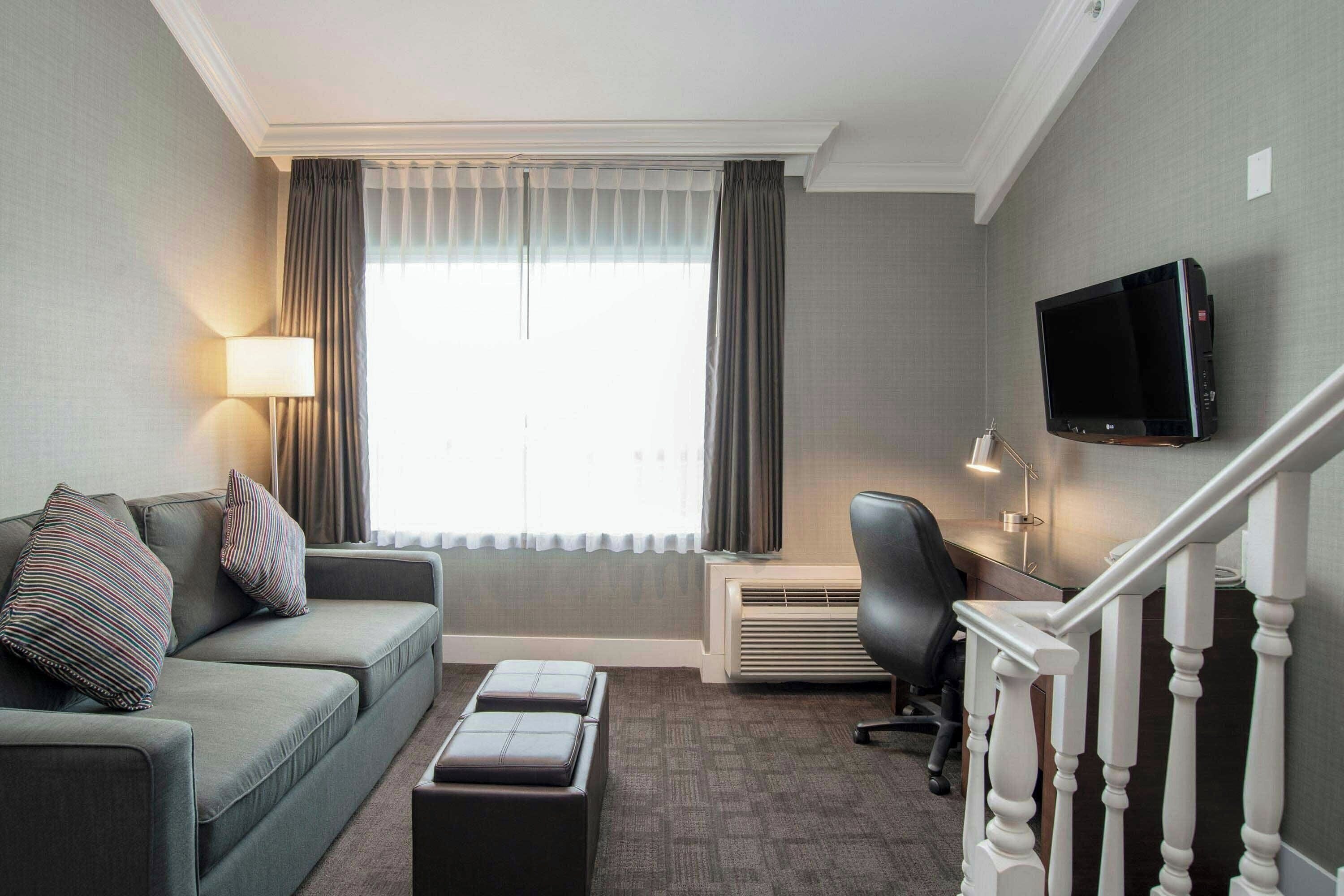 A hotel room with a sofa, a flat-screen TV mounted on the wall, a desk with a chair, and a staircase.