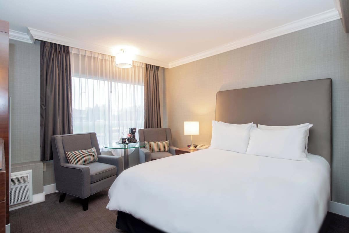 A hotel room with a large bed, a chair, a small table with a lamp, and a window with curtains.
