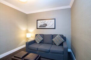 A modern hotel room with a grey sofa, two striped pillows, a framed artwork on the wall, and a small table with a lamp.