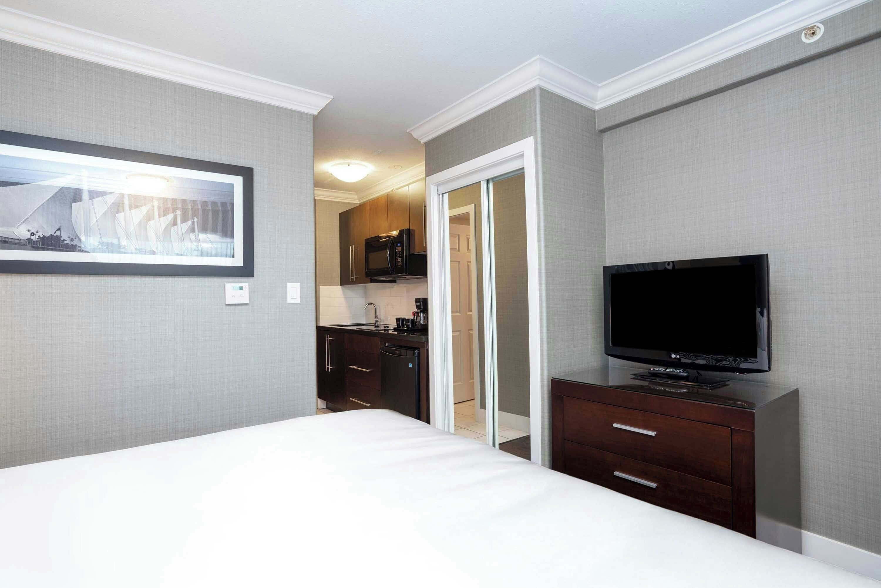 Una habitación de hotel con cama, televisor, una kitchenette pequeña con microondas y una puerta que da a otra habitación.