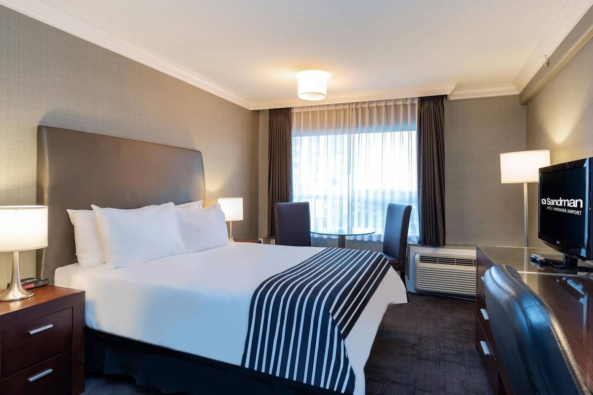 A hotel room with a large bed, a desk with a television, a chair, and an air conditioning unit.